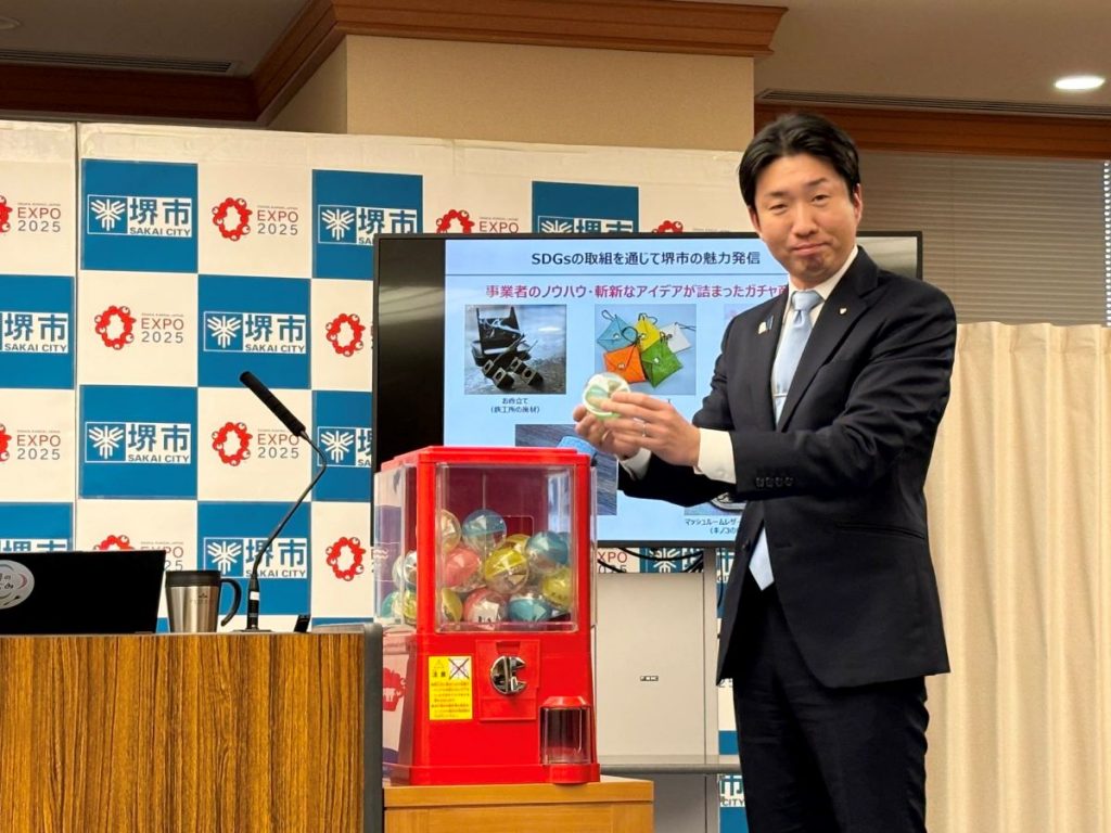 Mayor Nagafuji holding a gachapon capsule and standing next to a gachapon machine.